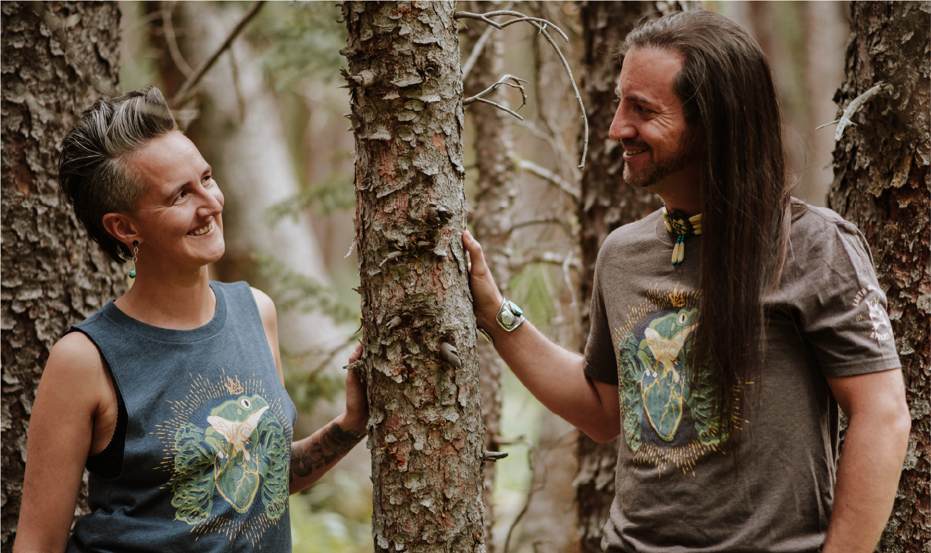 1 person wearing a blue tank and 1 wearing a brown shirt with a frog design, standing in the woods and smiling at each other.
