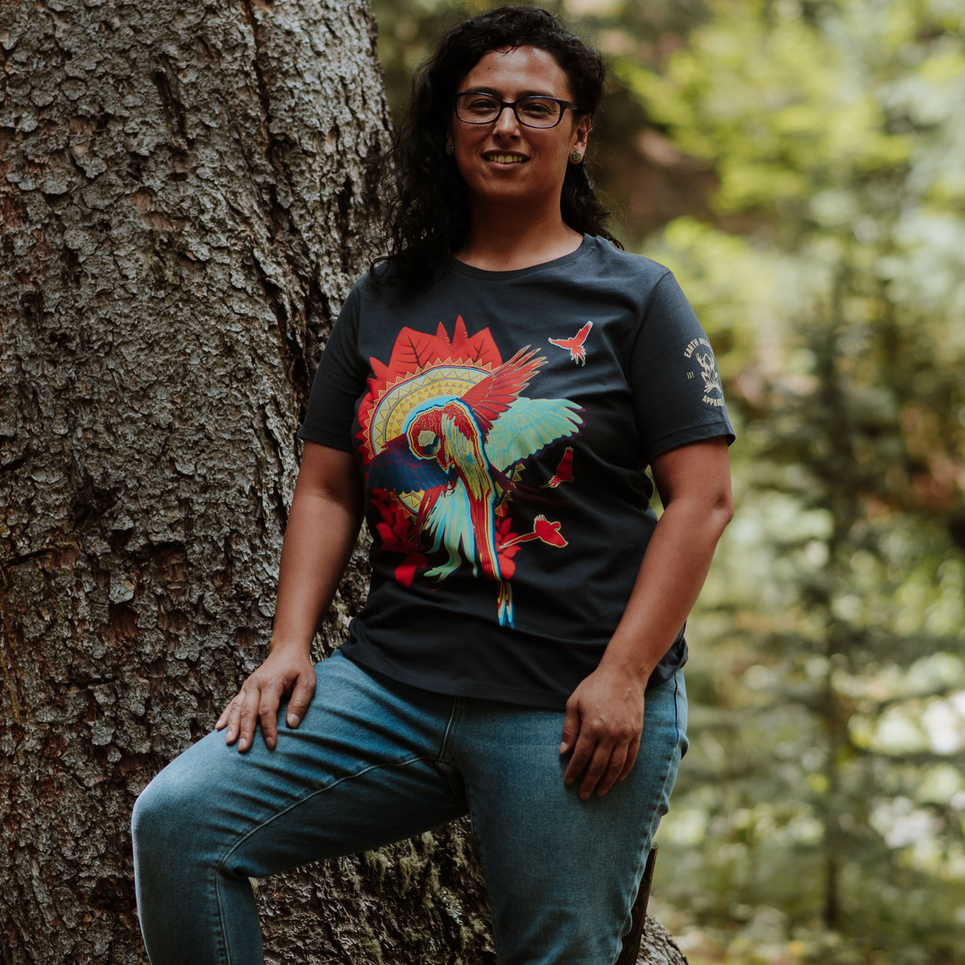 Woman wearing a dark grey t-shirt with a colorful macaw design, standing next to a tree in a forest.