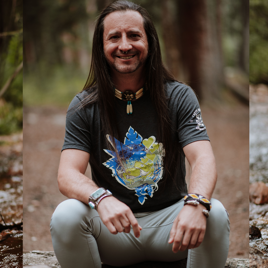 Non-binary person sitting outdoors in a forest wearing a black t-shirt with a colorful design.