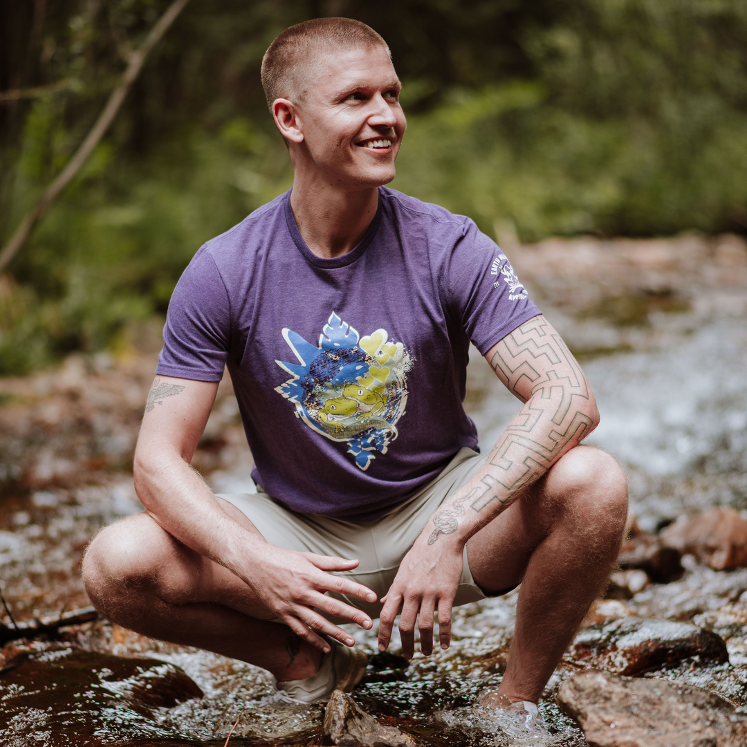 Man in a purple t-shirt with a colorful design sitting by a stream in a forest.
