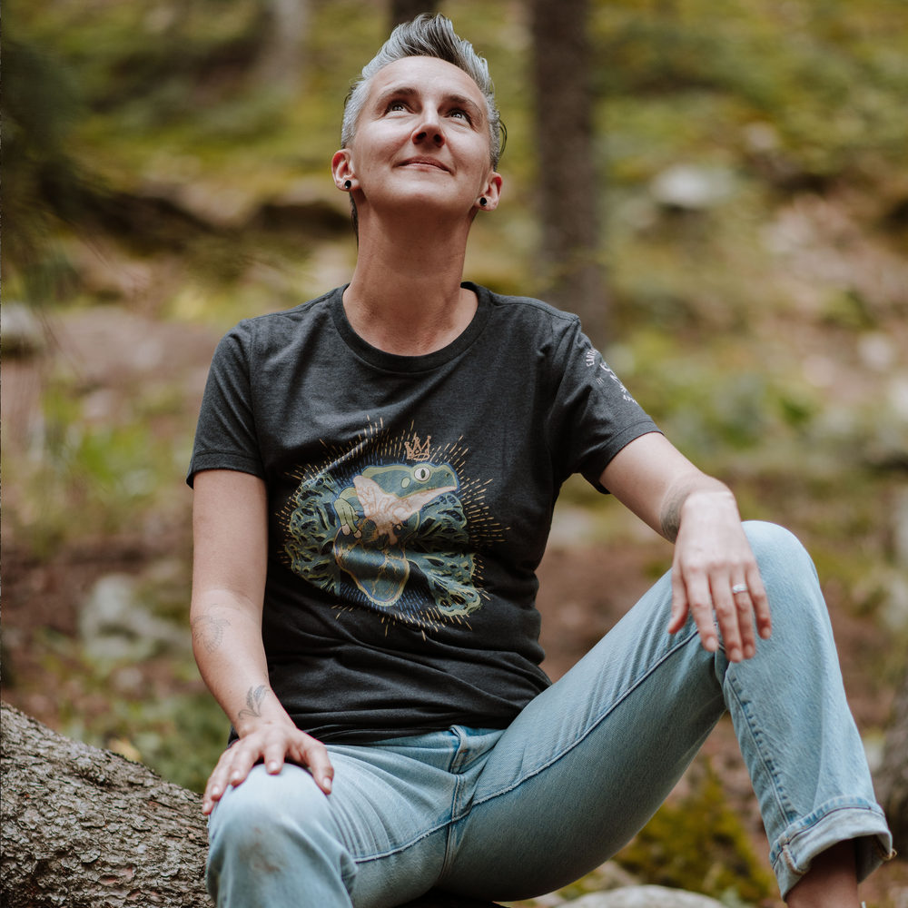 Woman sitting on a log in a forest wearing a black t-shirt with a frog graphic design and light blue jeans.