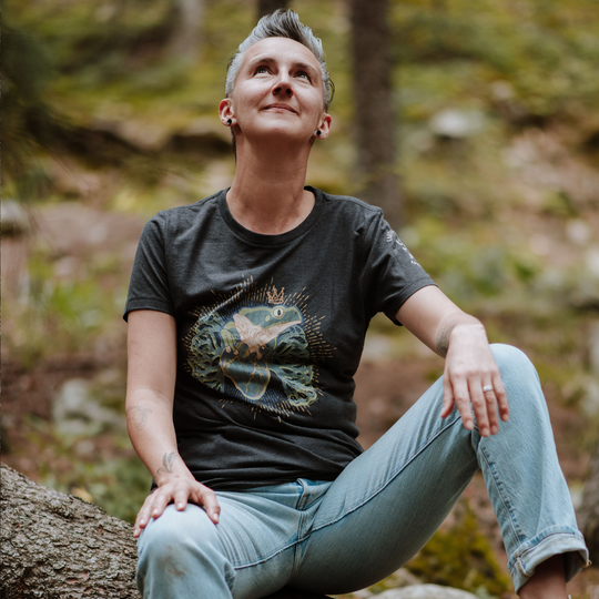 Woman sitting on a log in a forest wearing a black t-shirt with a frog graphic design and light blue jeans.