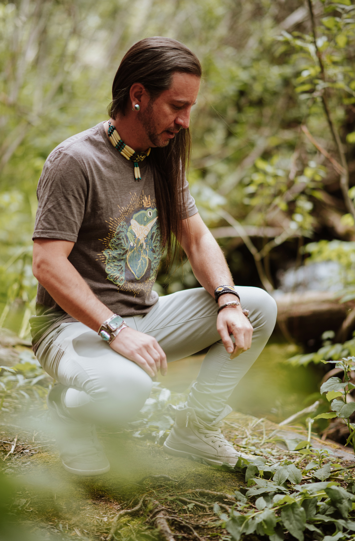Non-binary person sitting in a forest wearing a brown t-shirt and light pants.