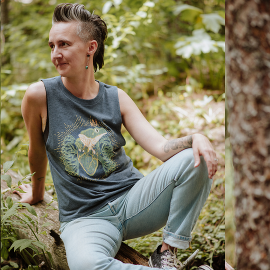 Woman sitting in a forest wearing a tank top with a graphic design.