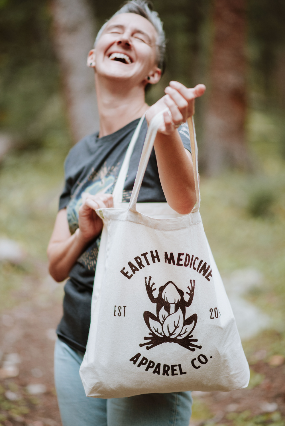 Person holding a tote bag with 'Earth Medicine Apparel Co.' branding in a forest setting