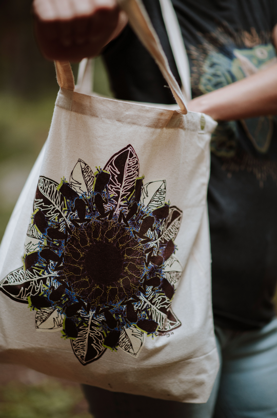Beige tote bag with leaf and frog mandala design held by a person outdoors