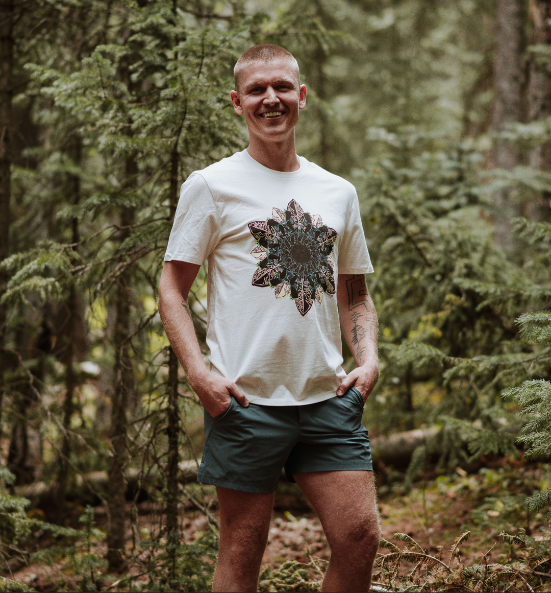 Man standing in a forest wearing a white t-shirt with a graphic design and green shorts.