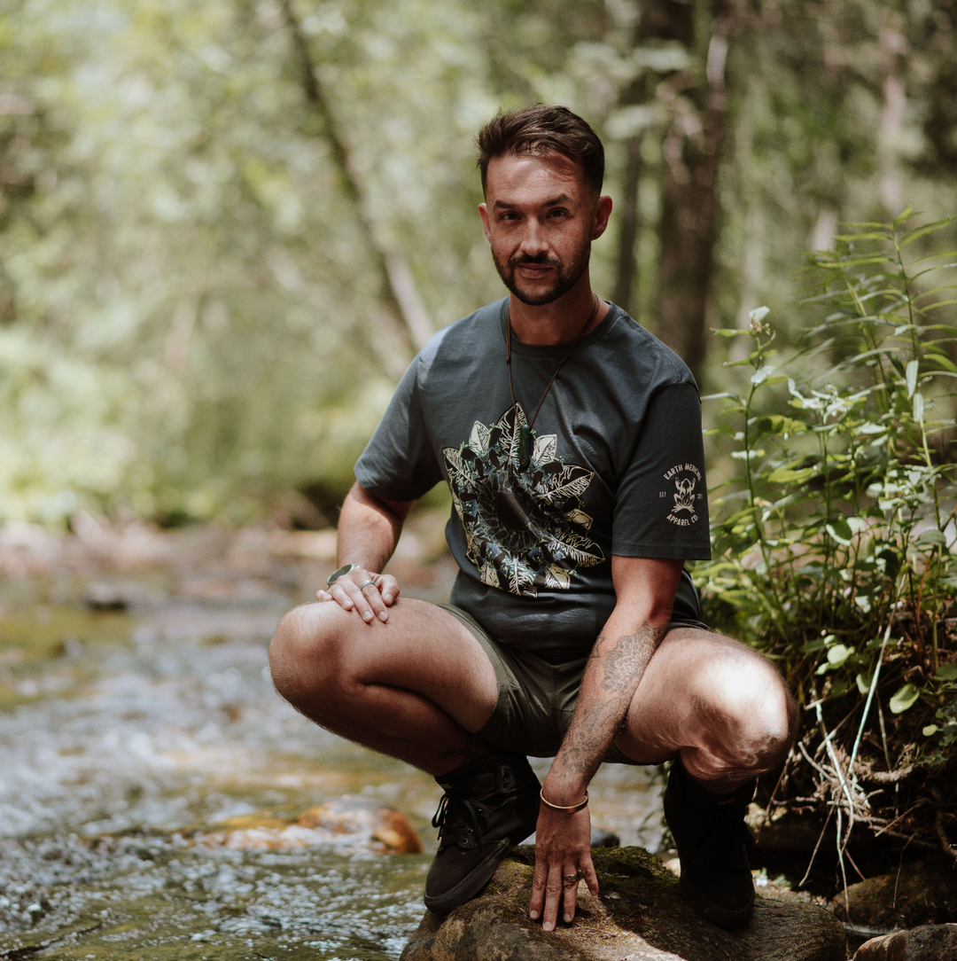 Man squatting by a stream in a forest in grey tshirt and green shorts.