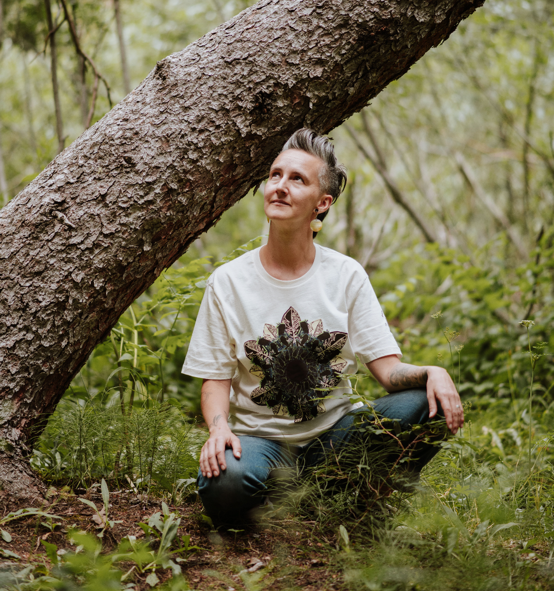 Woman sitting under a tree in a forest wearing a white t-shirt with a frog and leaf mandala design.