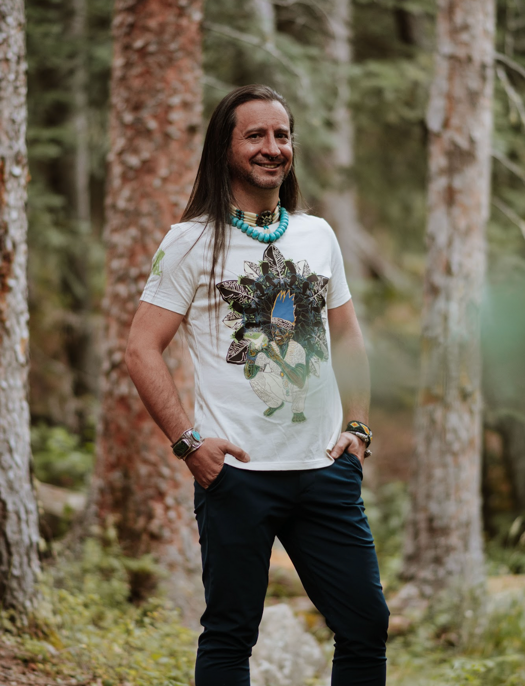 Non-binary person standing in a forest wearing a white t-shirt with a graphic design.