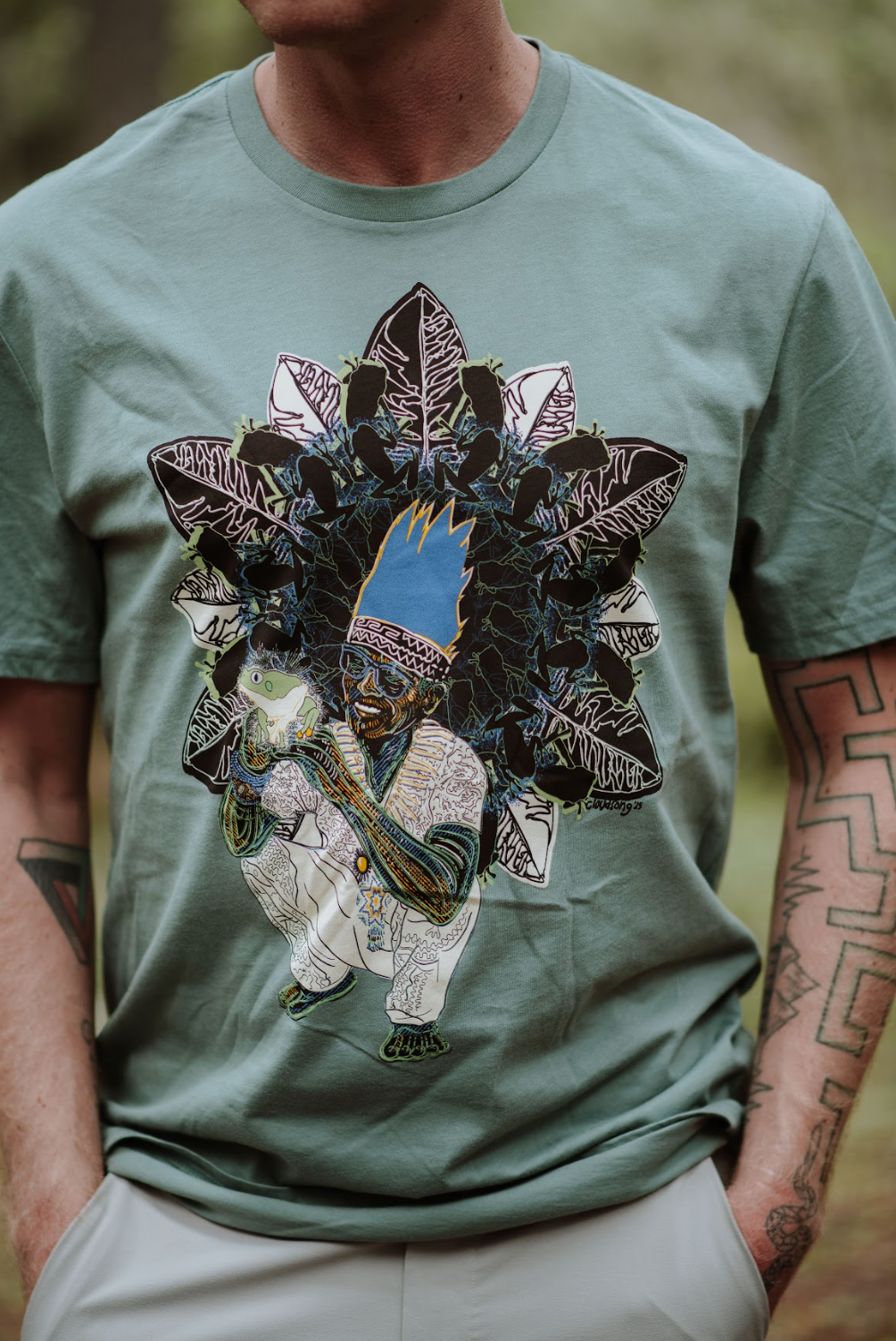 Person wearing a green t-shirt with a unique design of a Peruvian man, frog, and leaves.