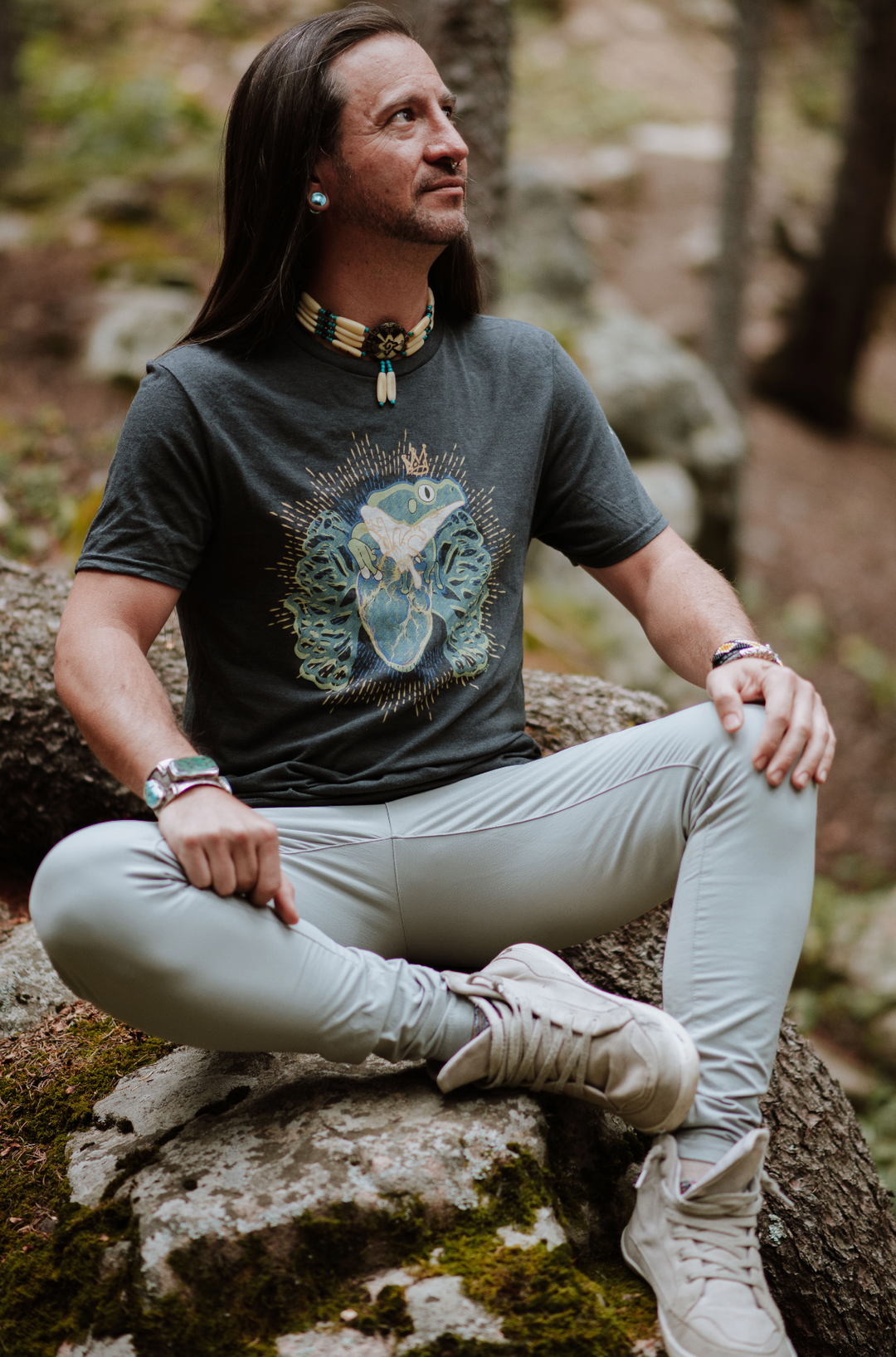 Non-binary person sitting on a rock in a natural setting wearing a graphic t-shirt and light pants.