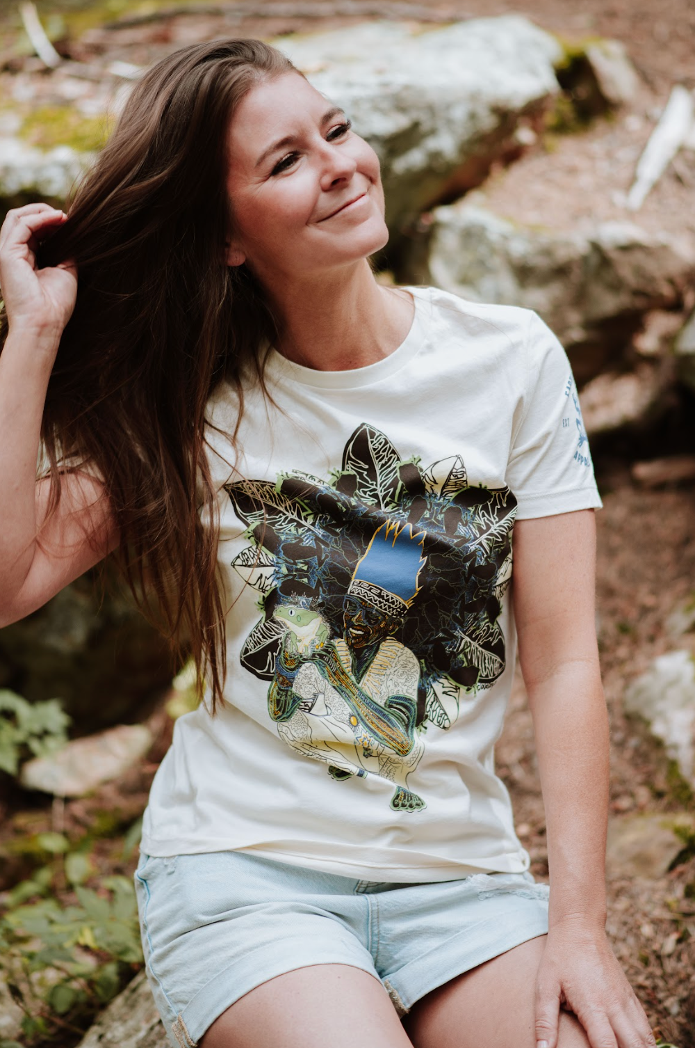 Woman wearing a white t-shirt with a graphic design, sitting outdoors on rocks.